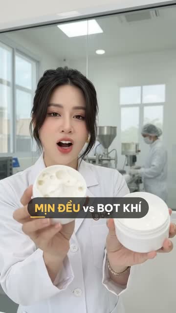 Chuyên viên BECO cầm hai hũ kem body, một hũ bề mặt lỗ chỗ bọt khí và một hũ mịn đều, phía sau vách kính là máy chiết rót và nhân viên đội mũ trùm