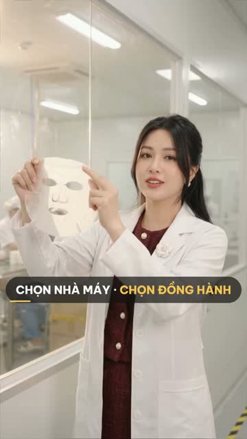 Chuyên viên BECO mặc áo blouse trắng giơ miếng mặt nạ lên trên hành lang vách kính của nhà xưởng