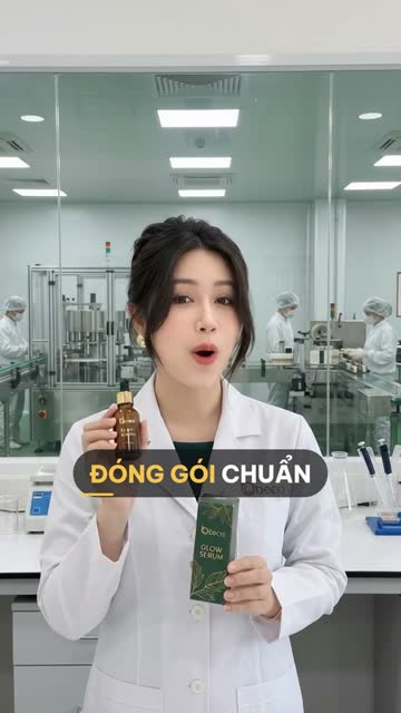 Chuyên viên BECO cầm chai serum màu hổ phách và hộp giấy xanh lá, phía sau vách kính là dây chuyền đóng gói với nhân viên đội mũ trùm