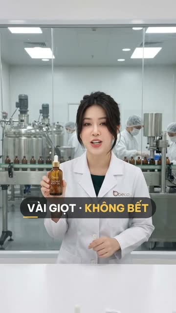 Chuyên viên BECO cầm chai dầu dưỡng thể thuỷ tinh màu hổ phách có ống nhỏ giọt, phía sau vách kính là bồn khuấy inox và hàng chai hổ phách trên băng chuyền