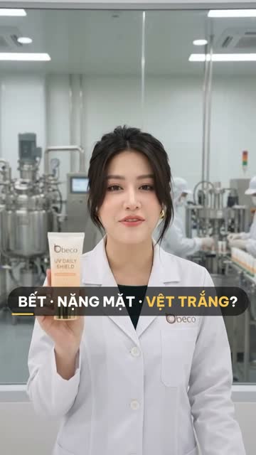 Chuyên viên BECO cầm tuýp kem chống nắng "UV Daily Shield", phía sau vách kính là bồn pha chế inox và công nhân đứng ở dây chuyền chiết tuýp