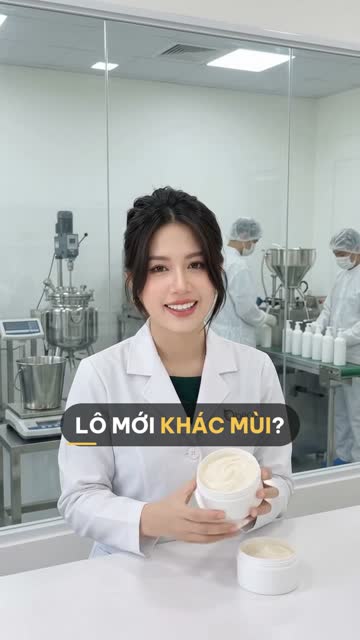 Chuyên viên BECO cầm hũ kem trắng đã mở nắp, bên cạnh là một hũ kem nhỏ, phía sau vách kính là nồi khuấy inox, cân điện tử và hàng chai vòi bơm trên băng chuyền