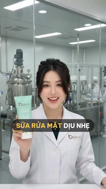Chuyên viên BECO cầm tuýp sữa rửa mặt màu xanh bạc hà, phía sau vách kính là bồn khuấy inox và máy chiết có nhân viên đứng máy