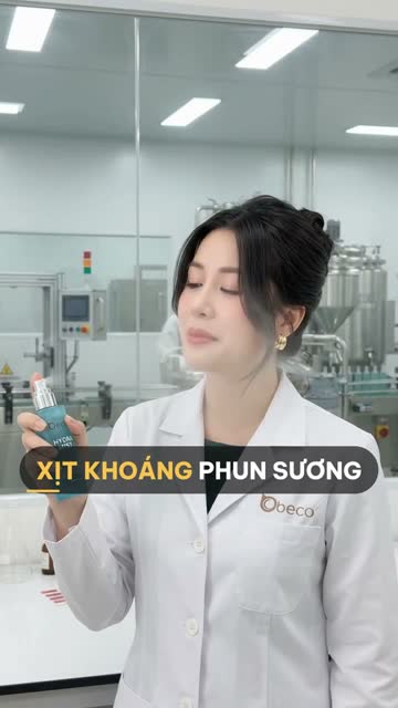 Chuyên viên BECO cầm chai xịt khoáng màu xanh ngọc, phía sau vách kính là máy chiết rót và bồn khuấy inox trong nhà xưởng
