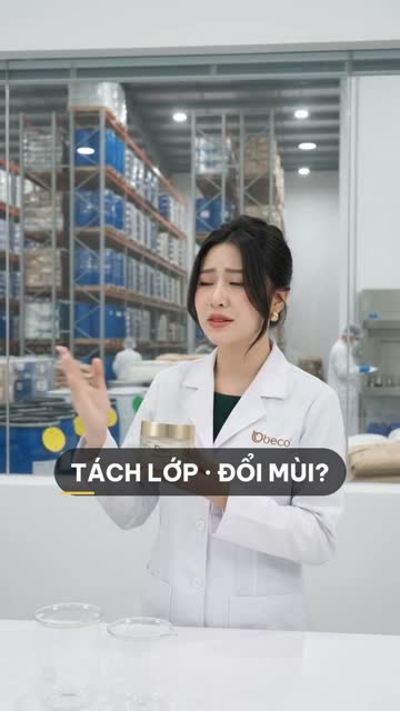 Chuyên viên BECO cầm hũ kem, phía sau vách kính là kho nguyên liệu với kệ thùng phuy xanh và bao hàng xếp nhiều tầng