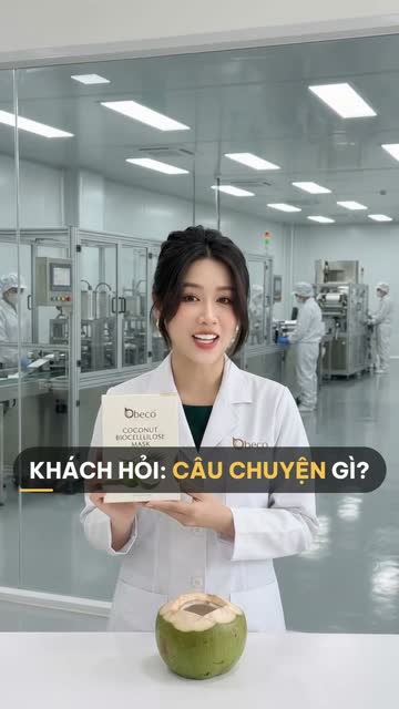 Chuyên viên BECO cầm hộp "Coconut Biocellulose Mask", trên bàn có trái dừa xiêm đã khui, phía sau vách kính là dây chuyền đóng gói mặt nạ