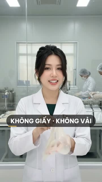 Chuyên viên BECO cầm miếng mặt nạ biocellulose trong mờ, phía sau vách kính là bồn inox và nhân viên đội mũ trùm đang đóng gói mặt nạ