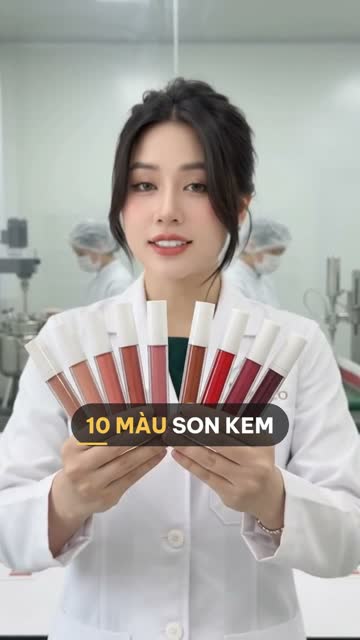 Chuyên viên BECO cầm xòe một bộ tuýp son kem từ tông nude đến đỏ mận, phía sau là khu sản xuất với nhân viên đội mũ trùm